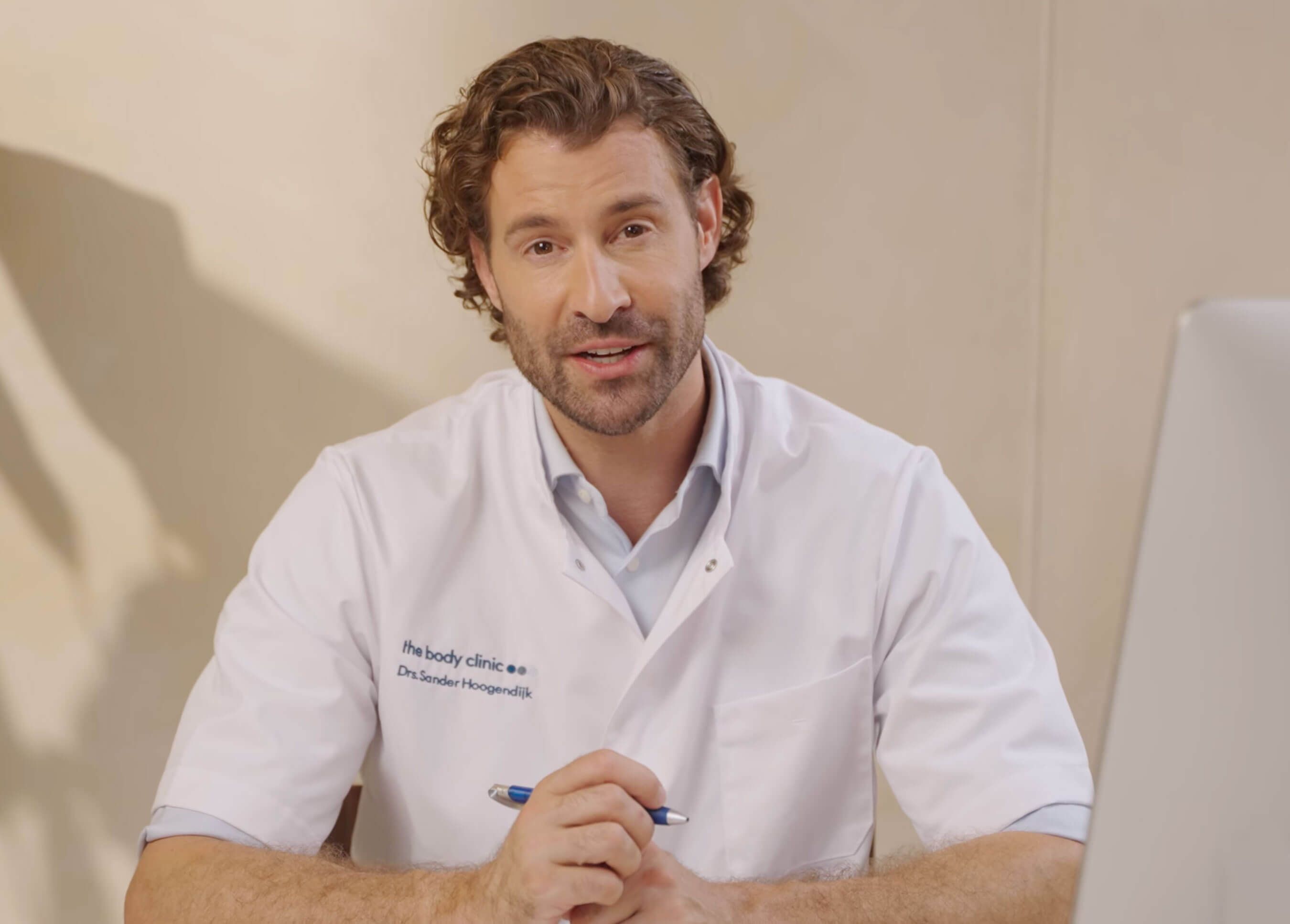 A male doctor in a white coat, with curly hair, speaks while holding a pen.