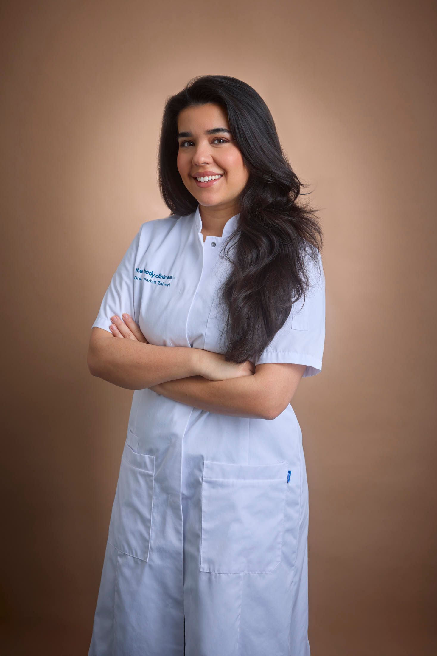 Smiling young woman in a white coat with arms crossed against a brown background.